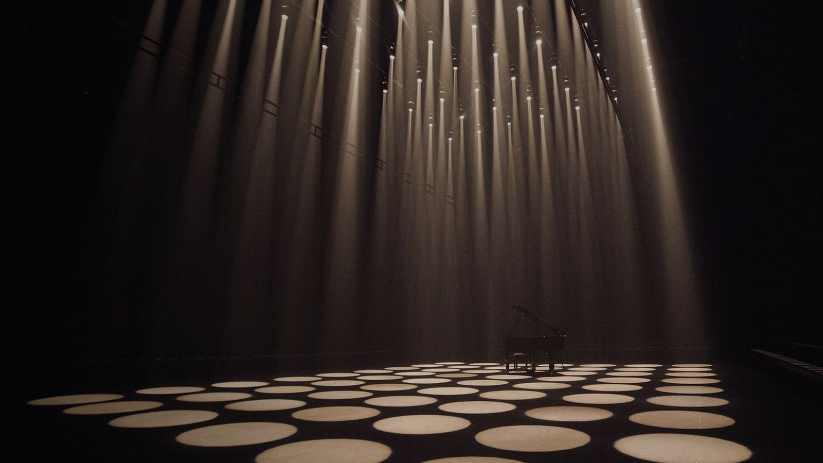 A grand piano lit by rows of beams on an empty stage